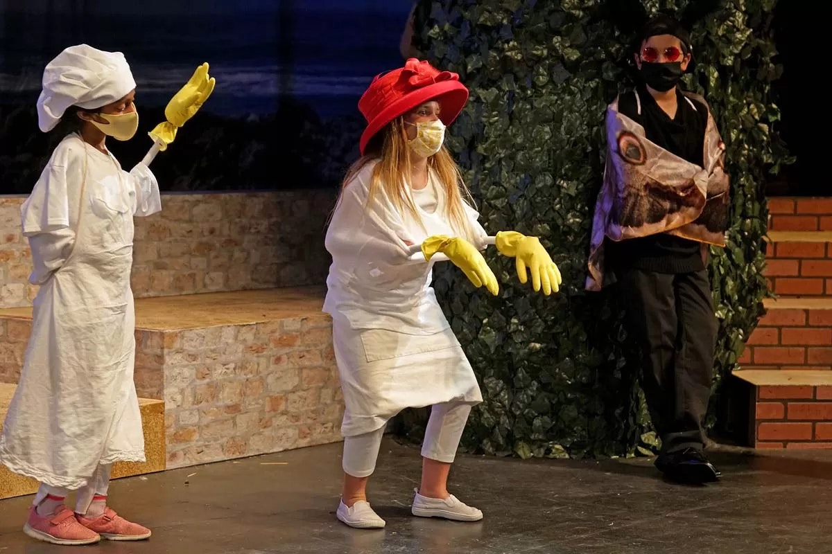 Children performing on stage in costumes with masks and gloves; one wears a red hat, another a chef's hat, and the third in dark attire.