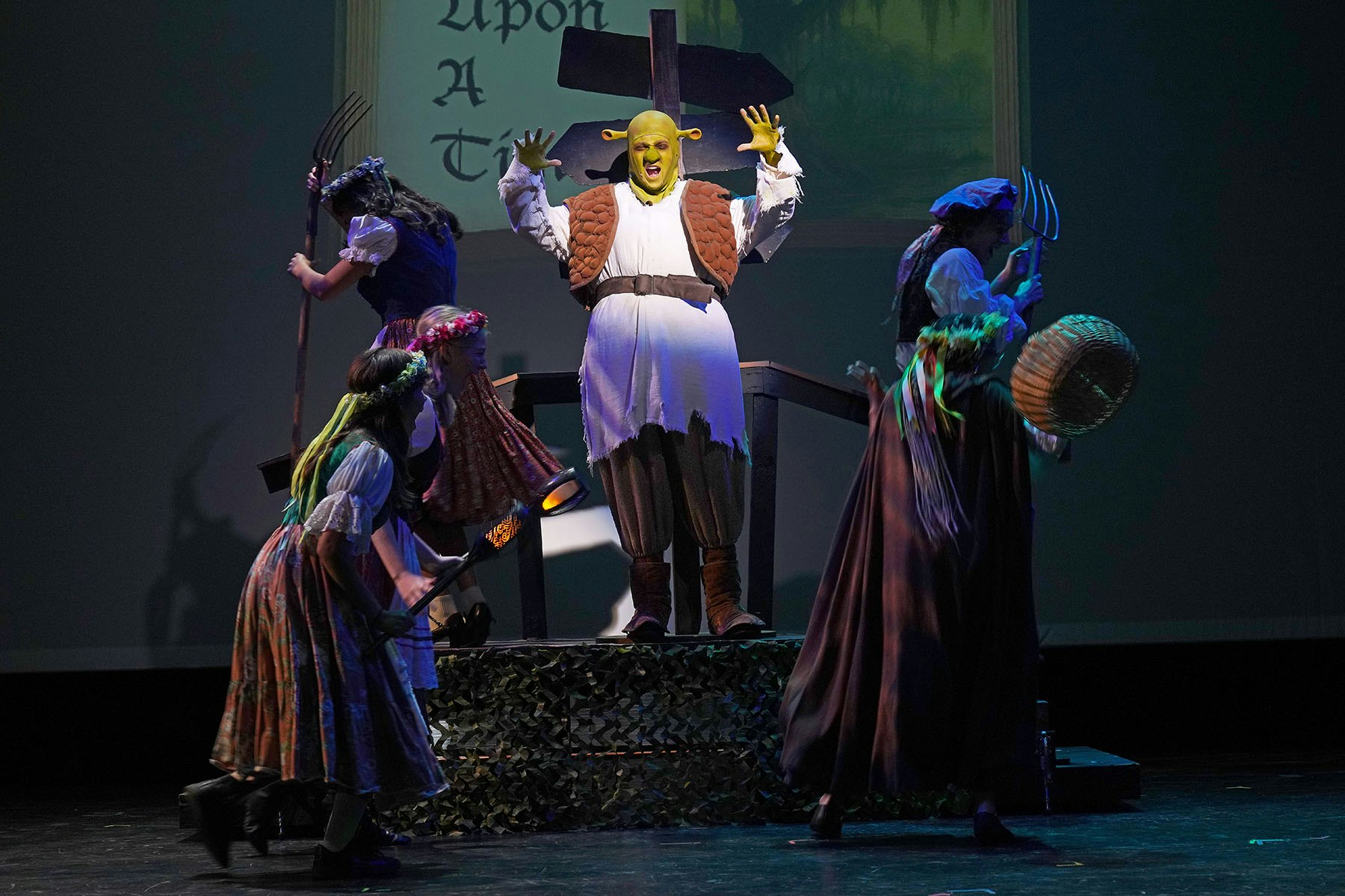 A person dressed as an ogre performs on stage with others in costume, set in a fairy tale theme.