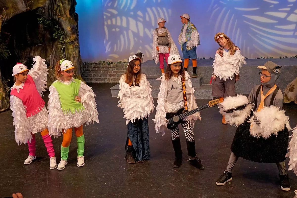 Children in bird costumes perform on stage, with some playing instruments and wearing colorful leggings.