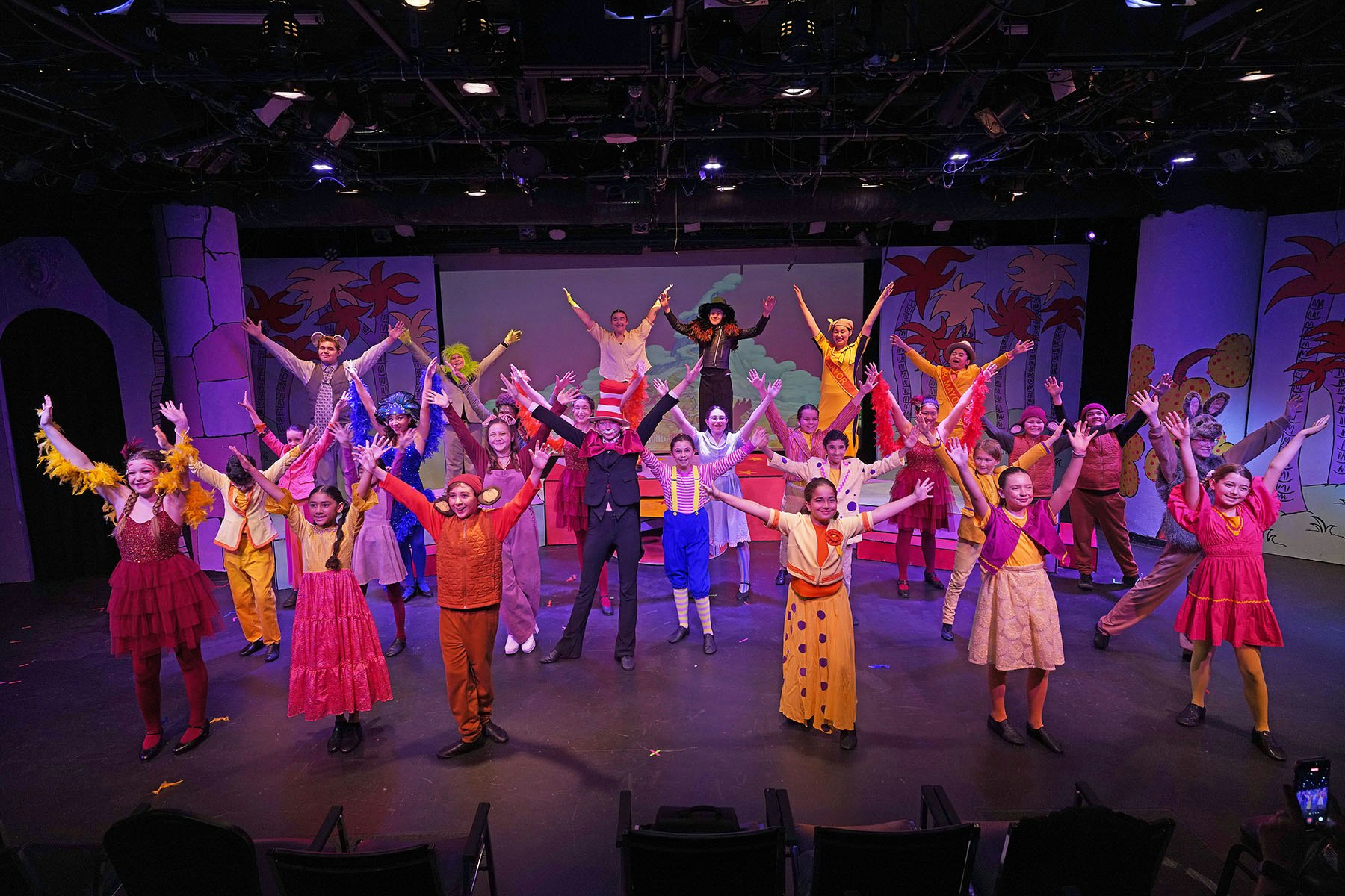 A vibrant theater performance featuring a diverse cast of children in colorful costumes, all joyfully posing on stage.