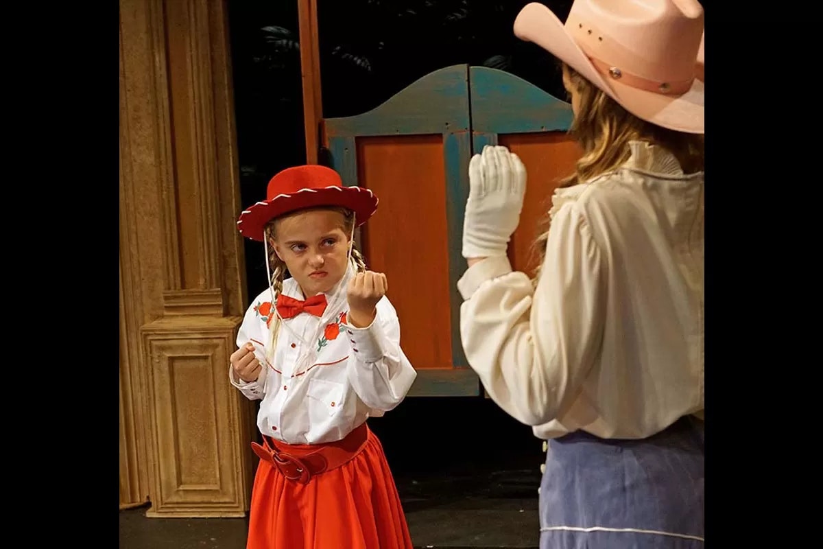 Two people dressed as cowboys face off, one with a fierce expression and fists raised, the other with a hand up.