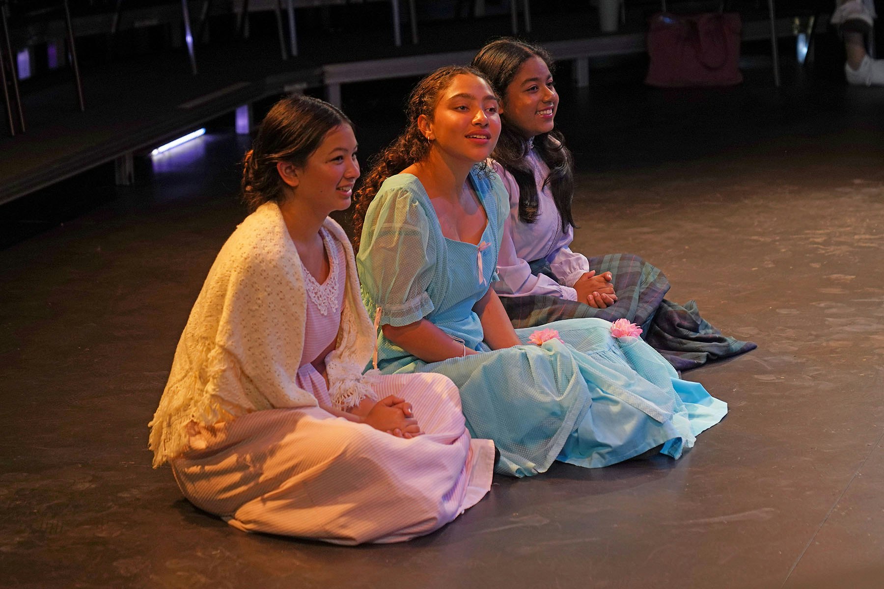 Three young girls in colorful period costumes sit smiling on a stage, engaged in a performance or rehearsal setting.
