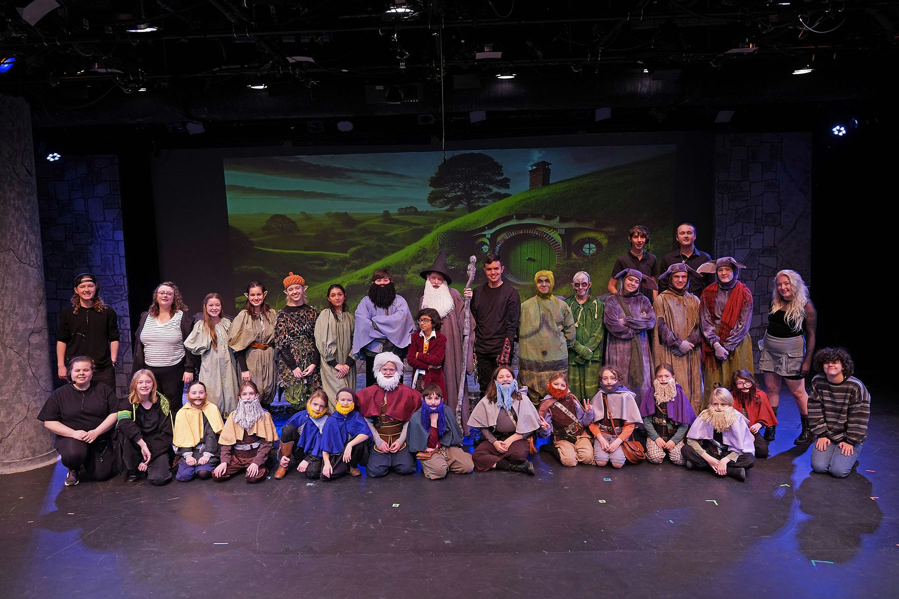 A group photo of people in costumes posing on stage with a Hobbit-themed backdrop.