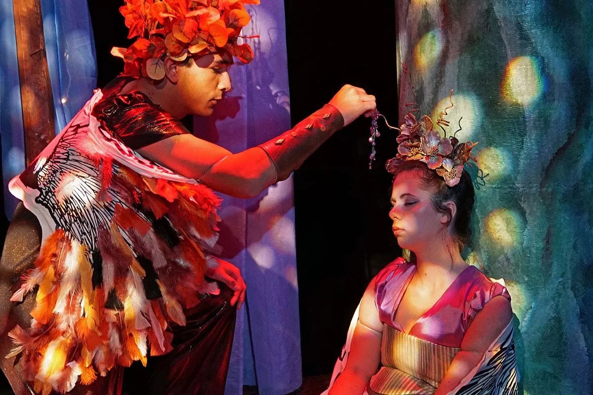 A person in a costume and flower headdress is sprinkling something over another person in a colorful setting.