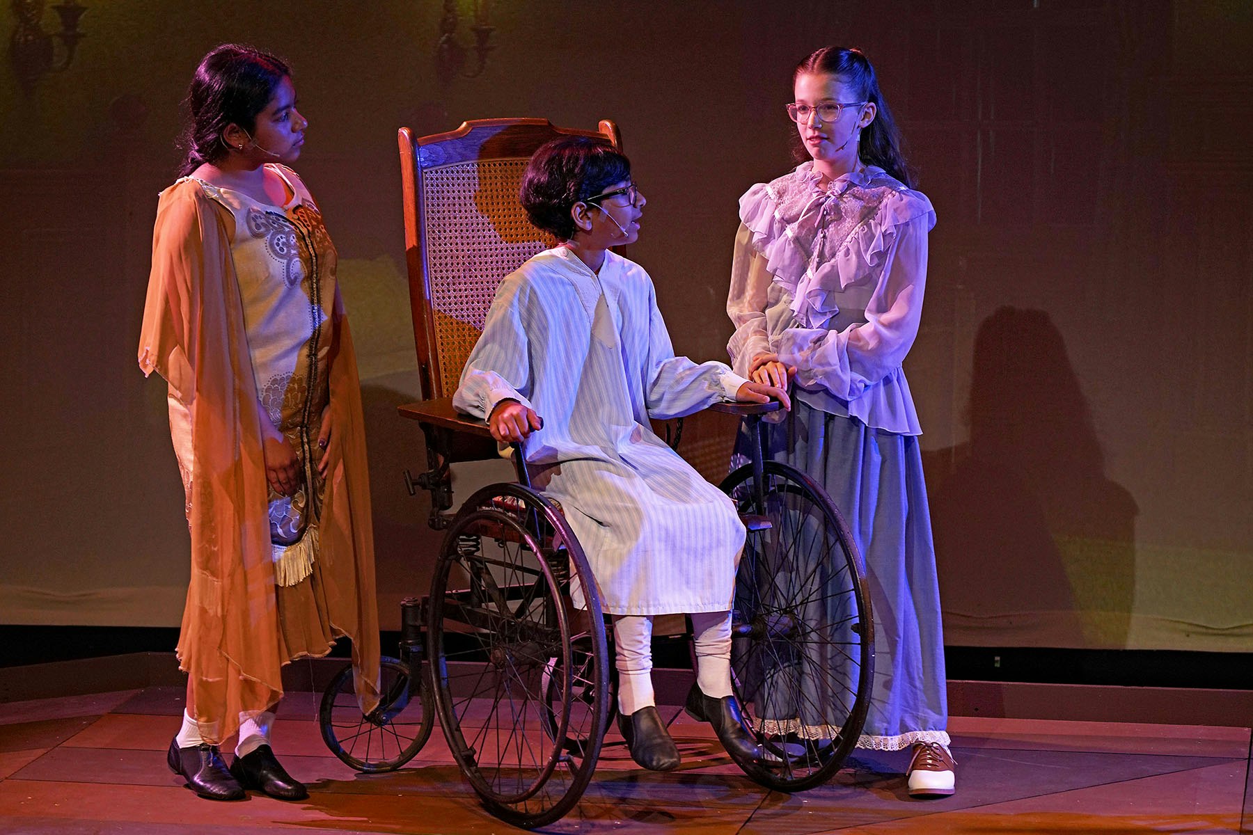 Three people on stage; a person sitting in a wheelchair, flanked by two others standing, wearing costumes suggestive of a historical play.