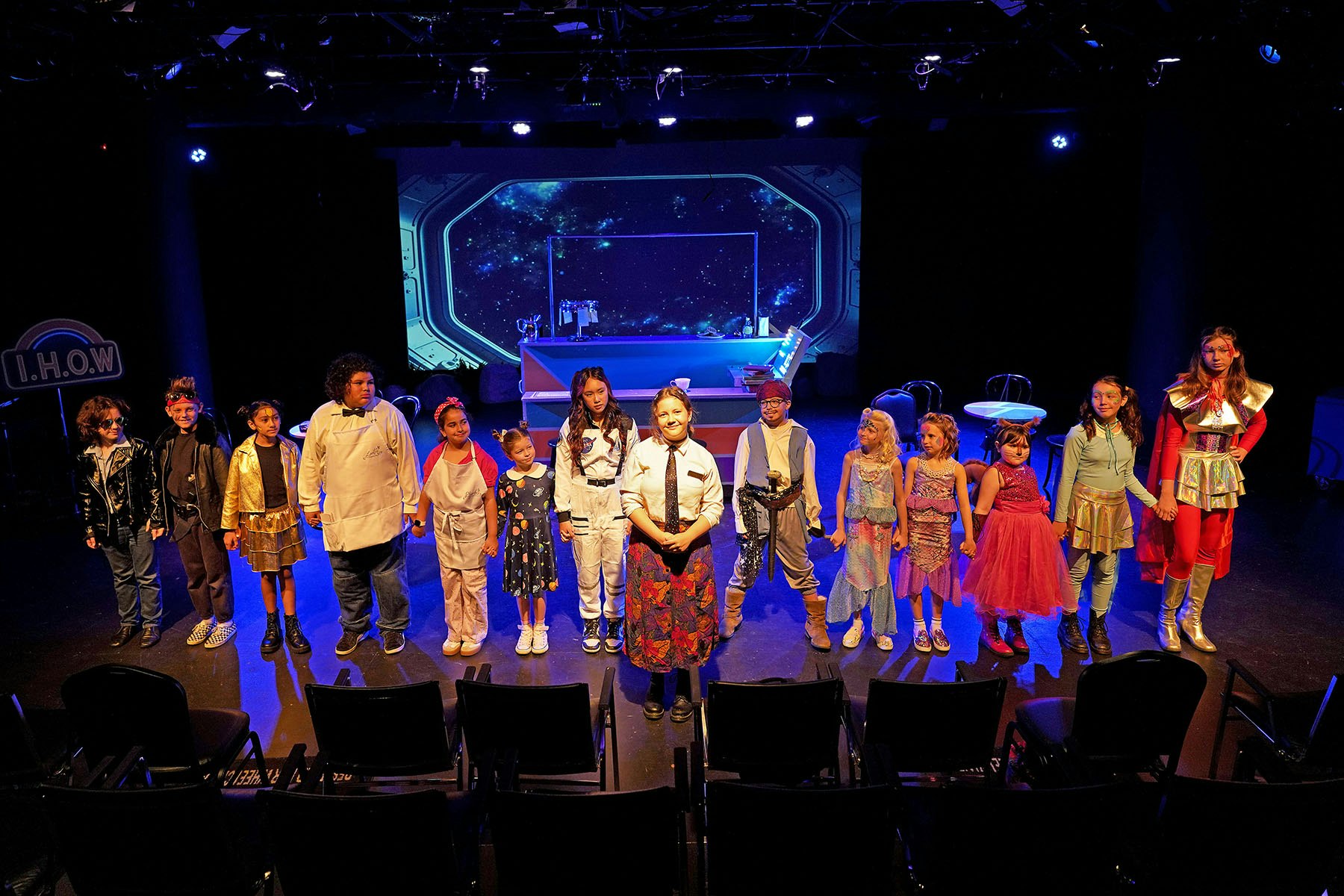A group of children in costumes on a stage with a space-themed background, holding hands.