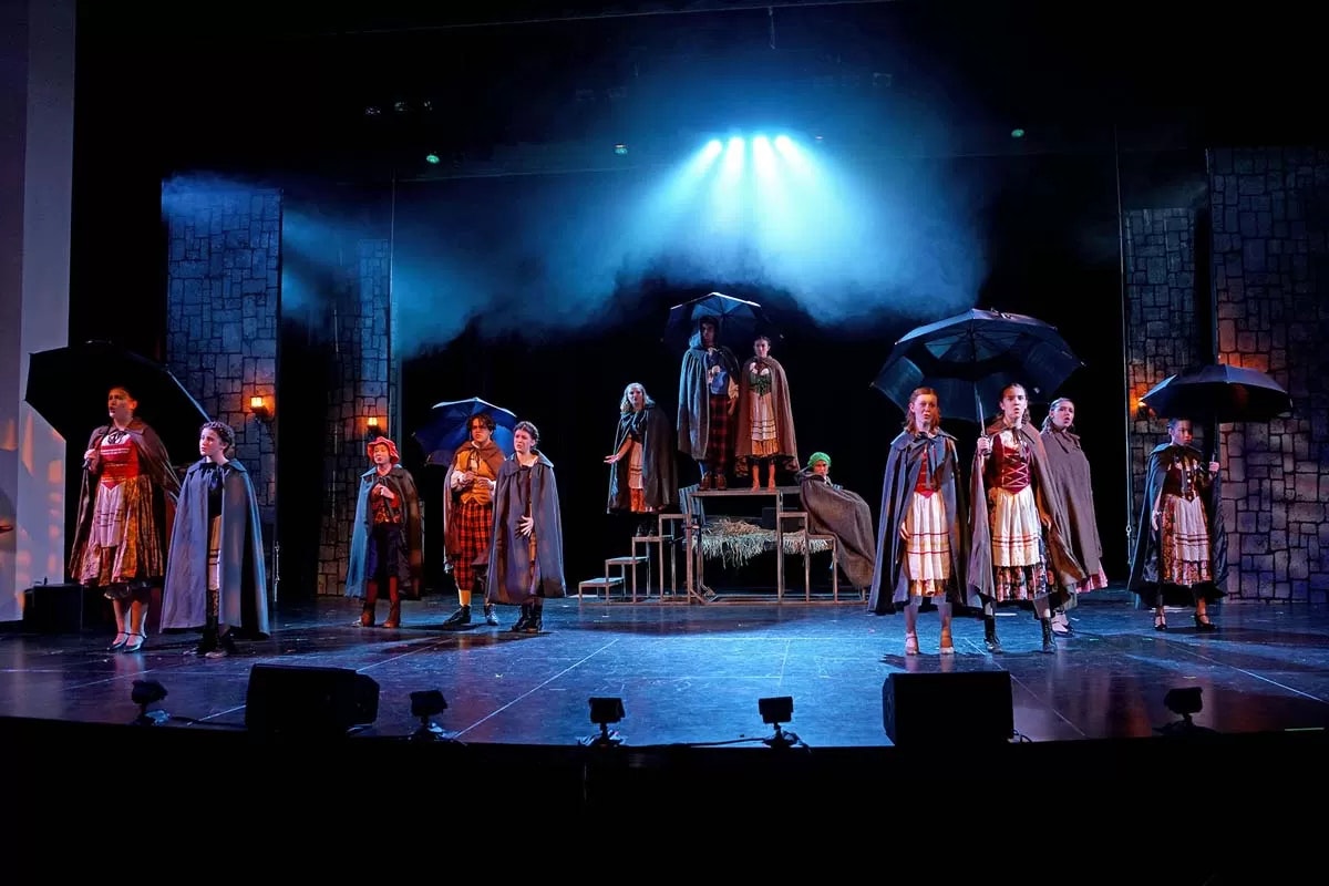 A theatrical stage scene with actors in costumes holding umbrellas, set against a moody, misty backdrop.