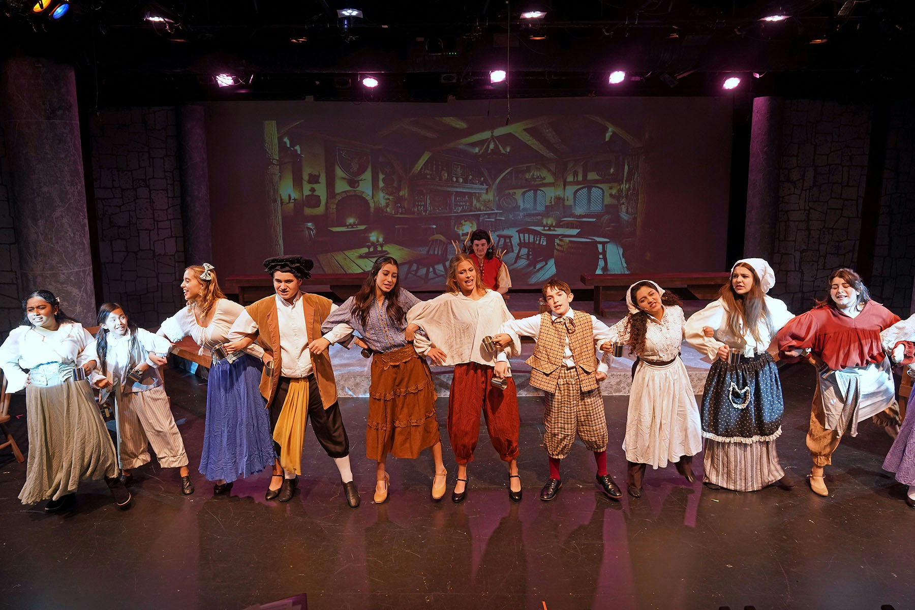 Performers in period costumes stand on stage with arms linked, vibrant lighting, and a backdrop of a vintage tavern interior.