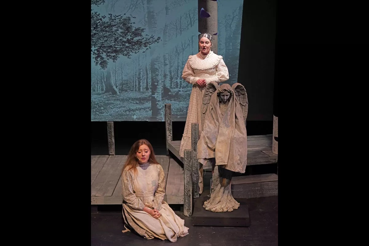 Two women in period costumes on stage, one standing, one seated; a statue and forest backdrop.