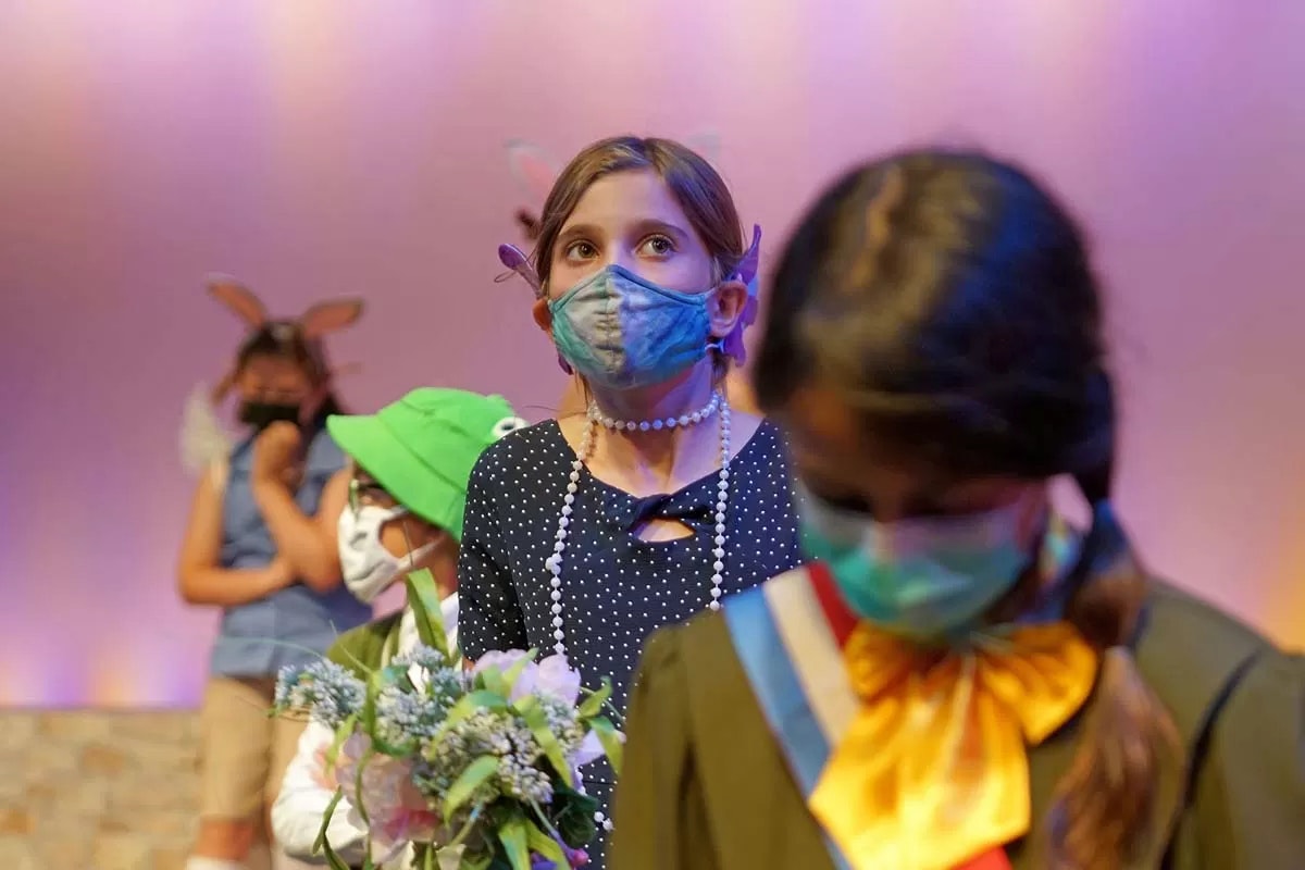 Children wearing costumes and masks, holding flowers, on a colorful stage.