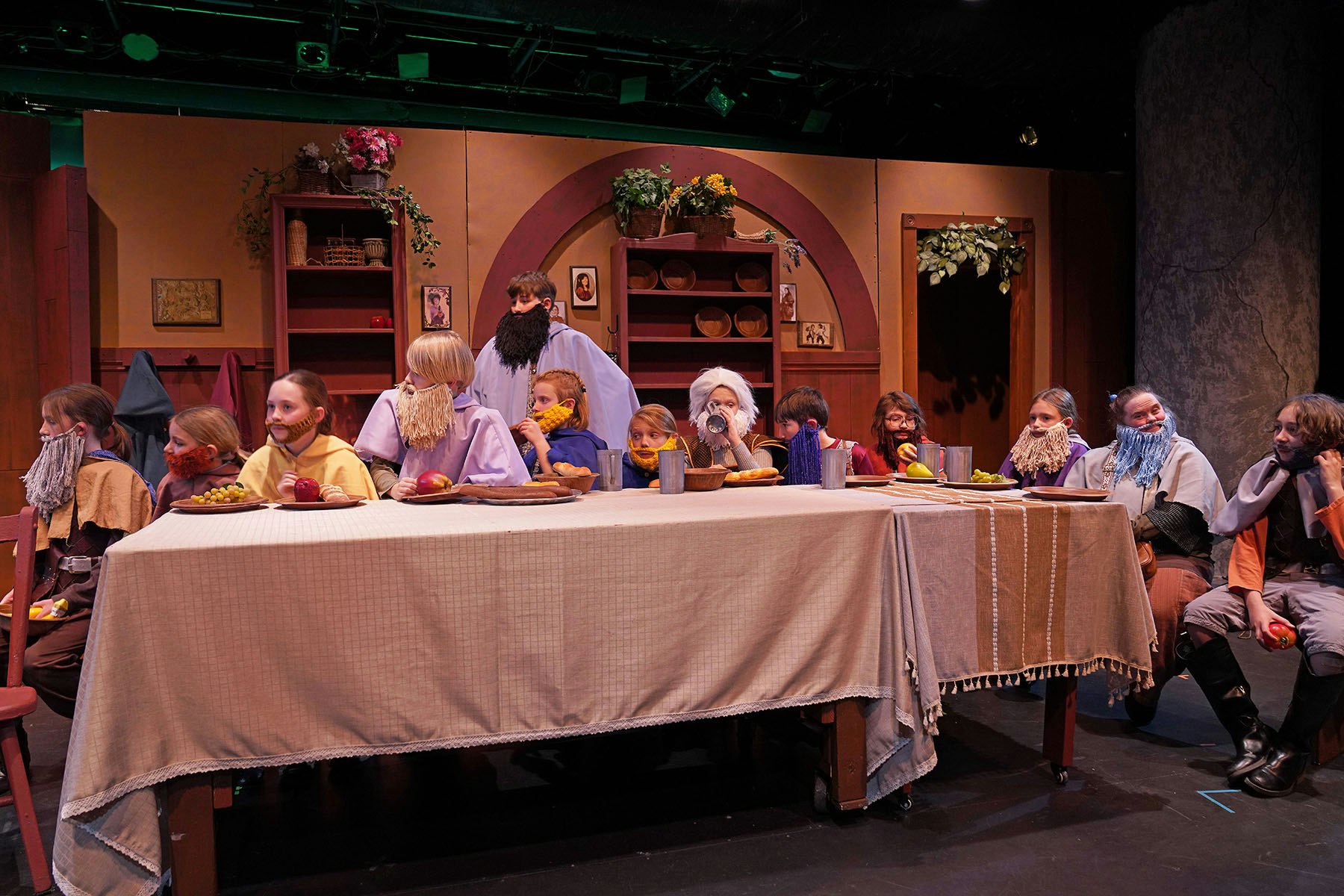 People in costumes with beards, sitting at a long table with food, possibly a theatrical performance.