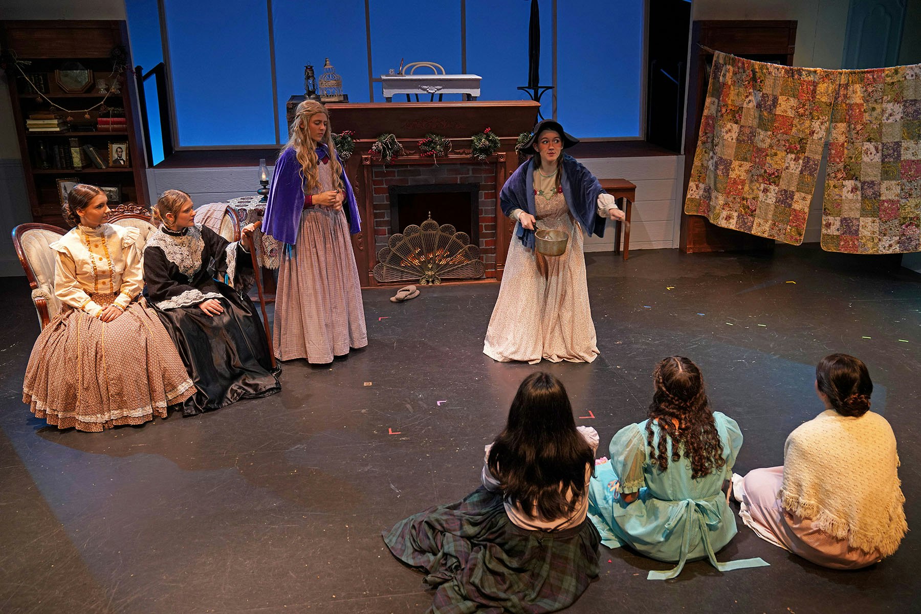 A stage performance featuring women in historical dresses, with one character acting out a scene while others watch.