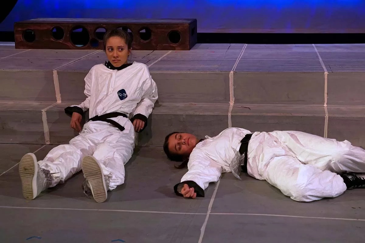 Two people in white suits; one sitting, one lying down, on stage steps.