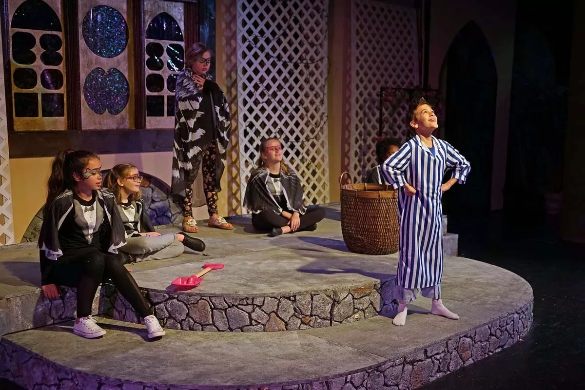 Children in costumes perform a play on stage with a colorful set and props.