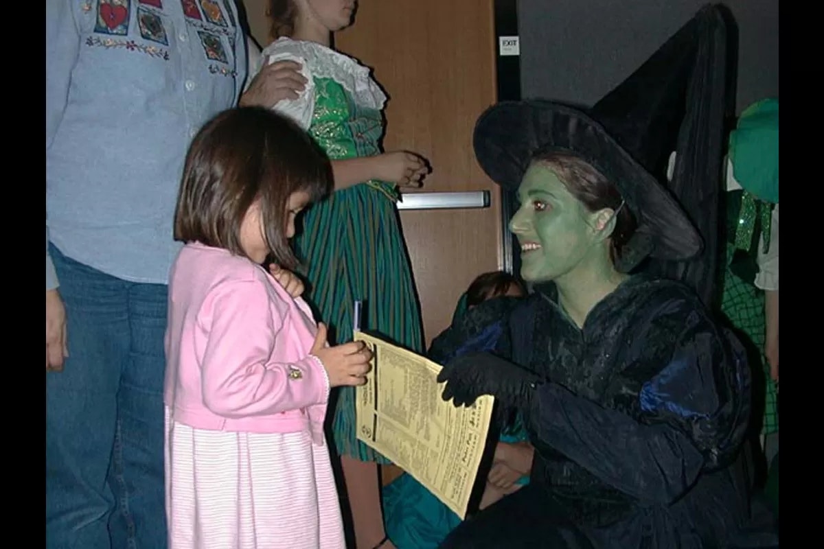 A woman dressed as a green-faced witch interacts with a young girl holding a paper.
