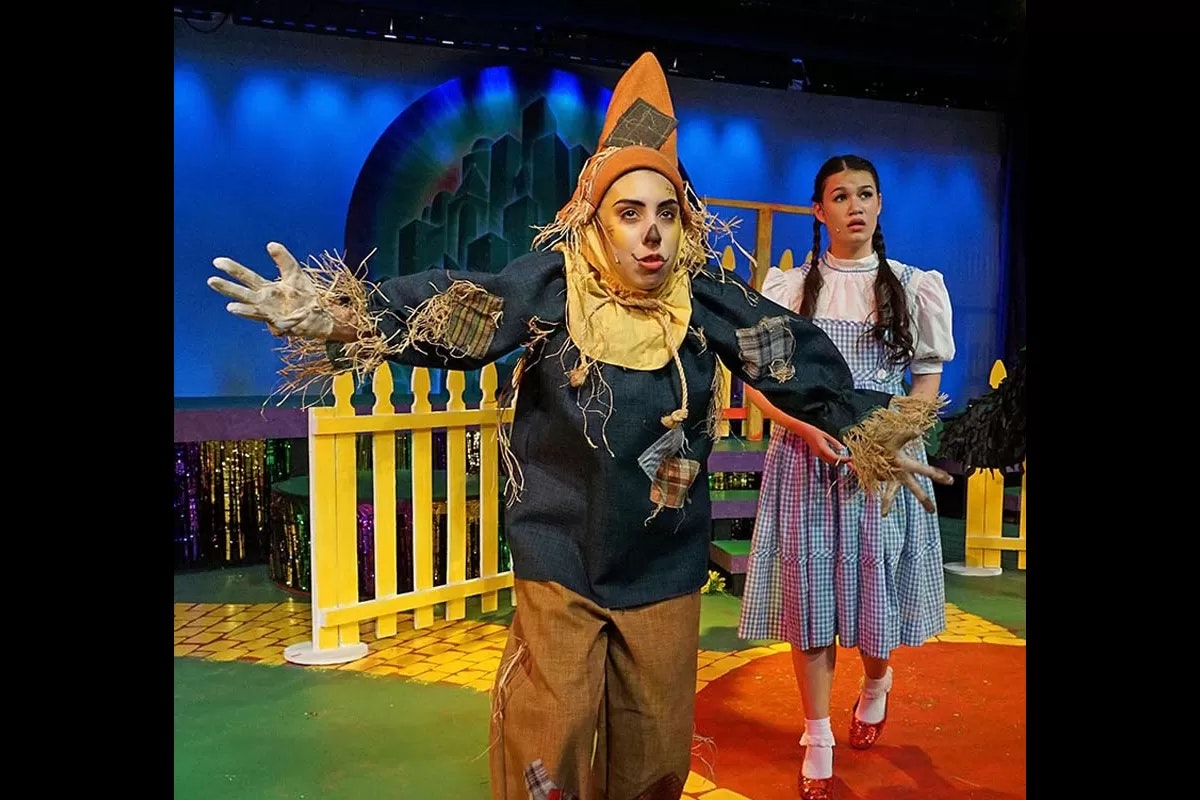A scarecrow and a girl in a gingham dress on stage, with a picket fence and colorful background.