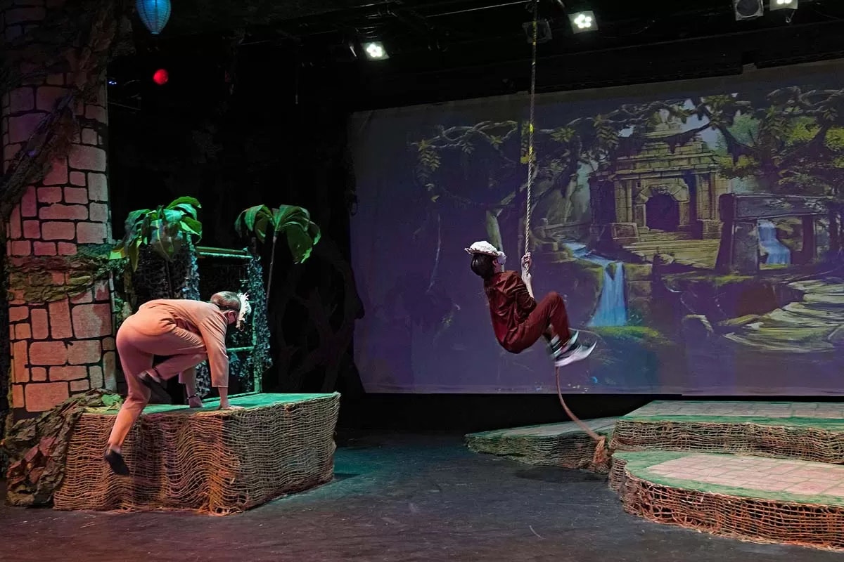 Two people in a stage play set in a jungle scene; one climbing, another swinging on a rope.