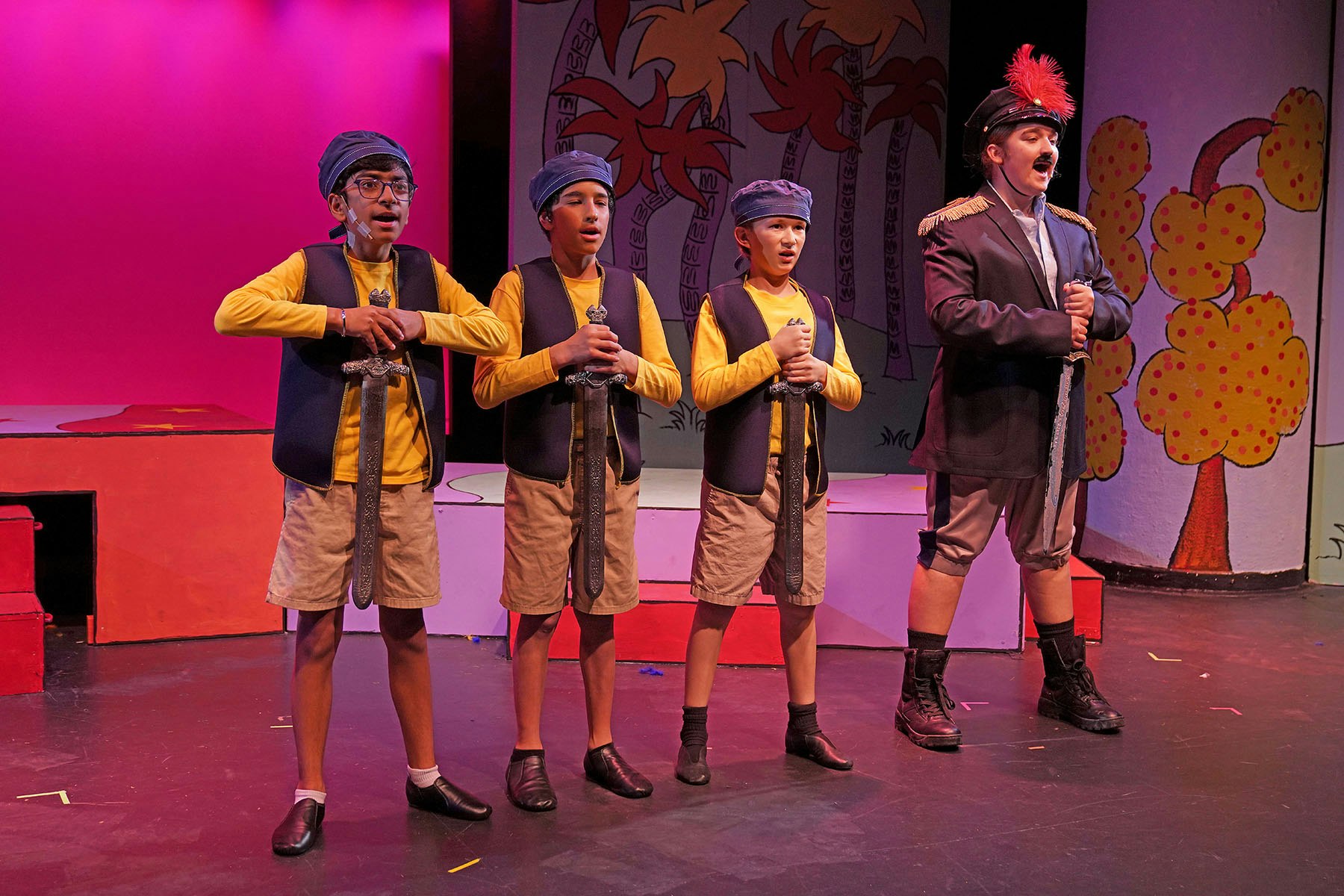 Four young performers in costume, holding swords on stage, with a colorful backdrop, likely from a theater production.