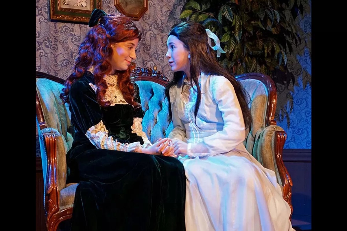 Two people in historical attire sit on a vintage sofa, holding hands, in a warmly lit room.