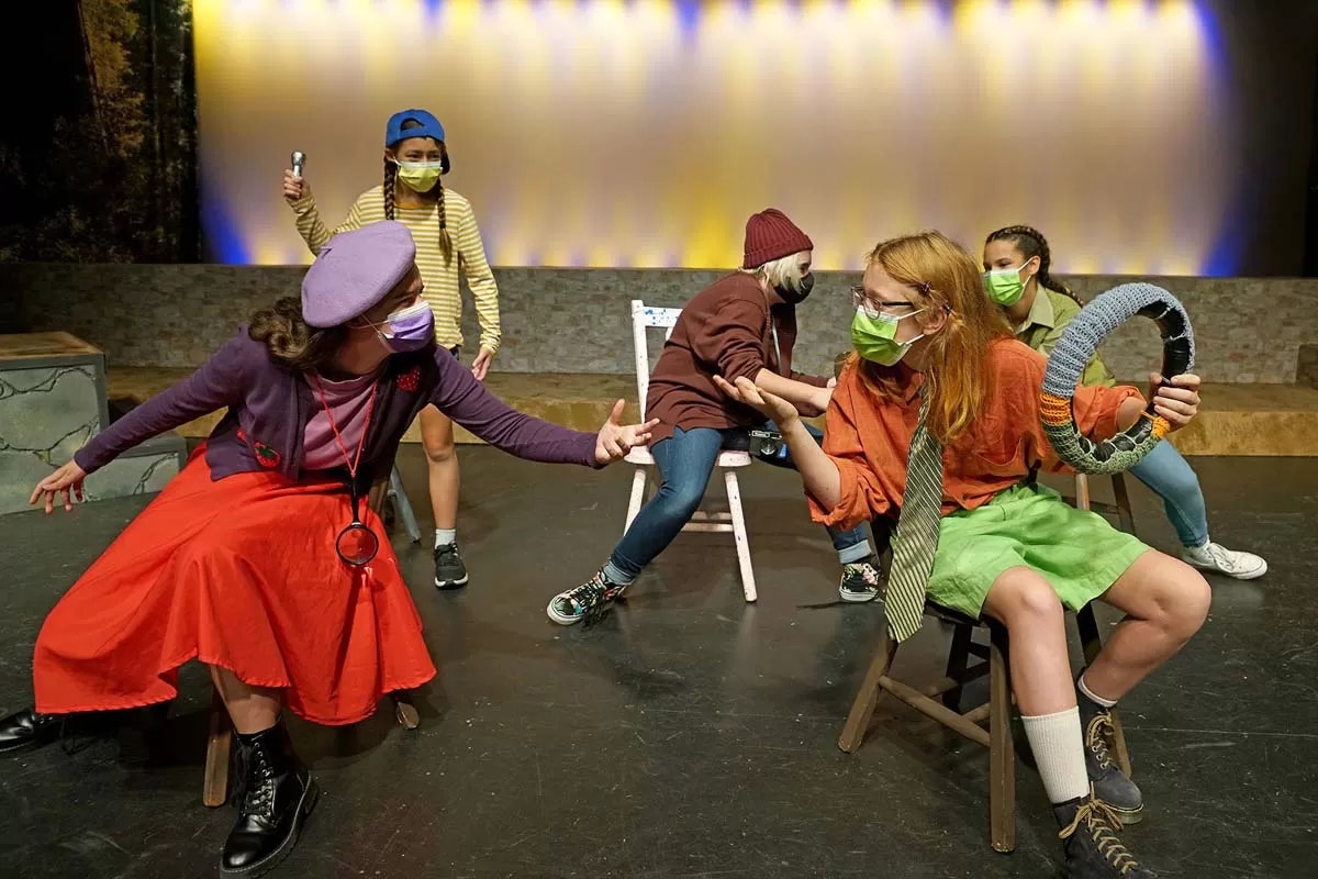 Five people in colorful costumes and masks perform on stage, featuring dynamic poses and theatrical expressions.