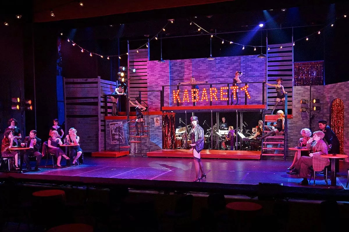 Cabaret-themed stage with performers, audience at tables, and musicians, under "KABARETT" sign.