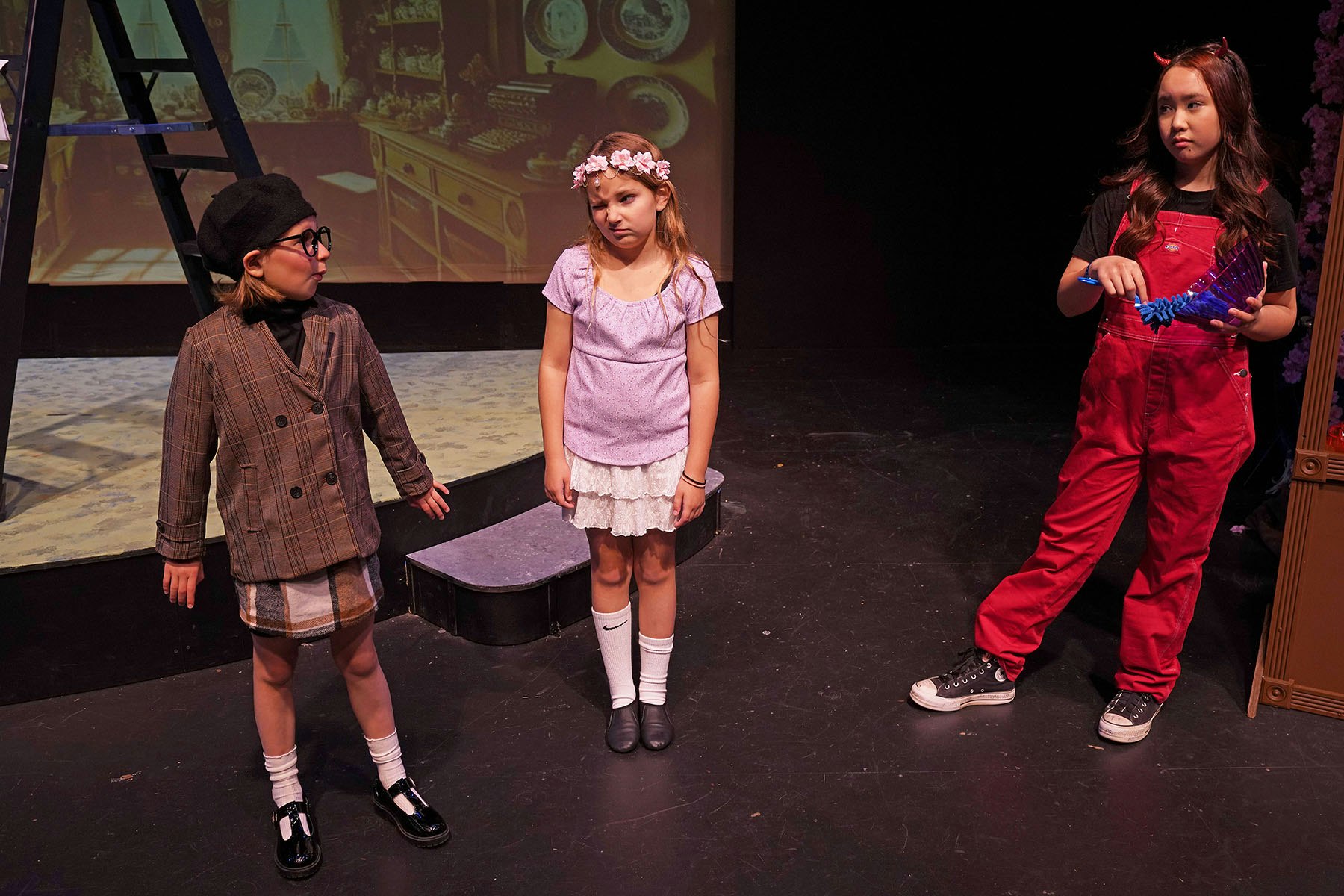 Three children in a play; one in plaid, another with flower headband, third with red overalls and devil horns, on a dimly lit stage.