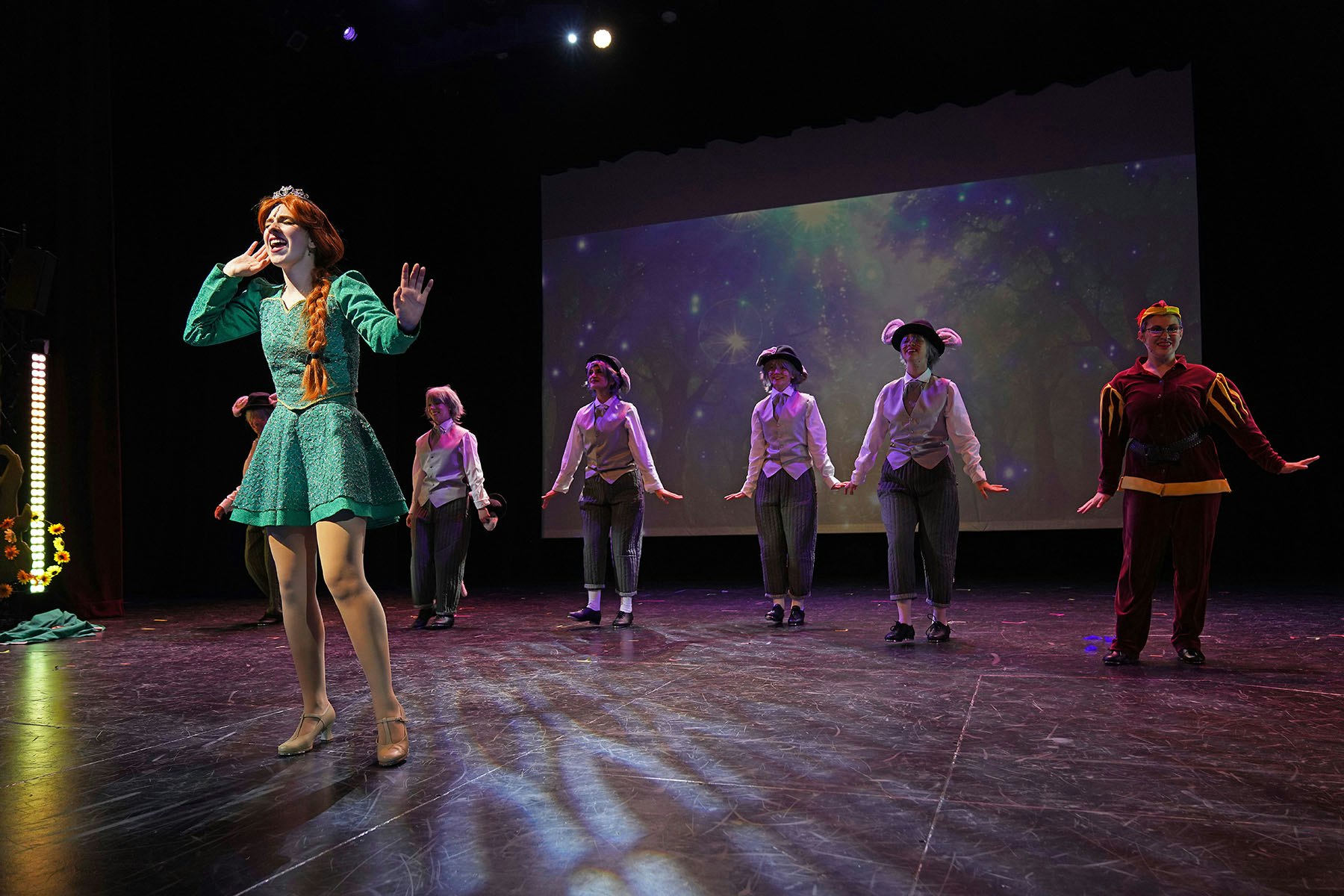 Performers on stage in colorful costumes, with a forest-themed backdrop.