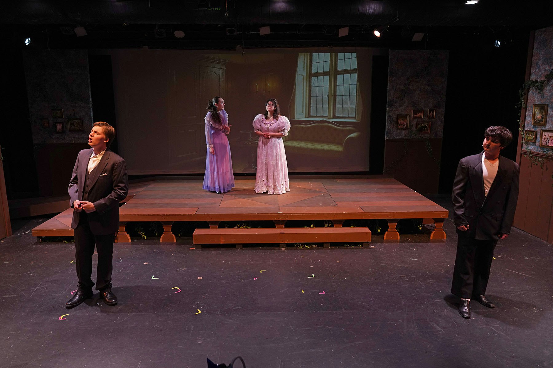 Four people on stage in a theater scene, two in dresses, two in suits, with a projected background of a room with a window.