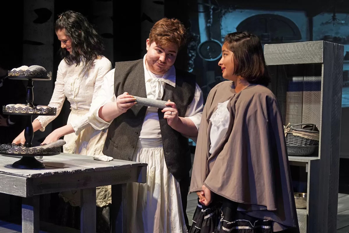 Three people in period costumes are acting in a theatrical scene with a table and baked goods.