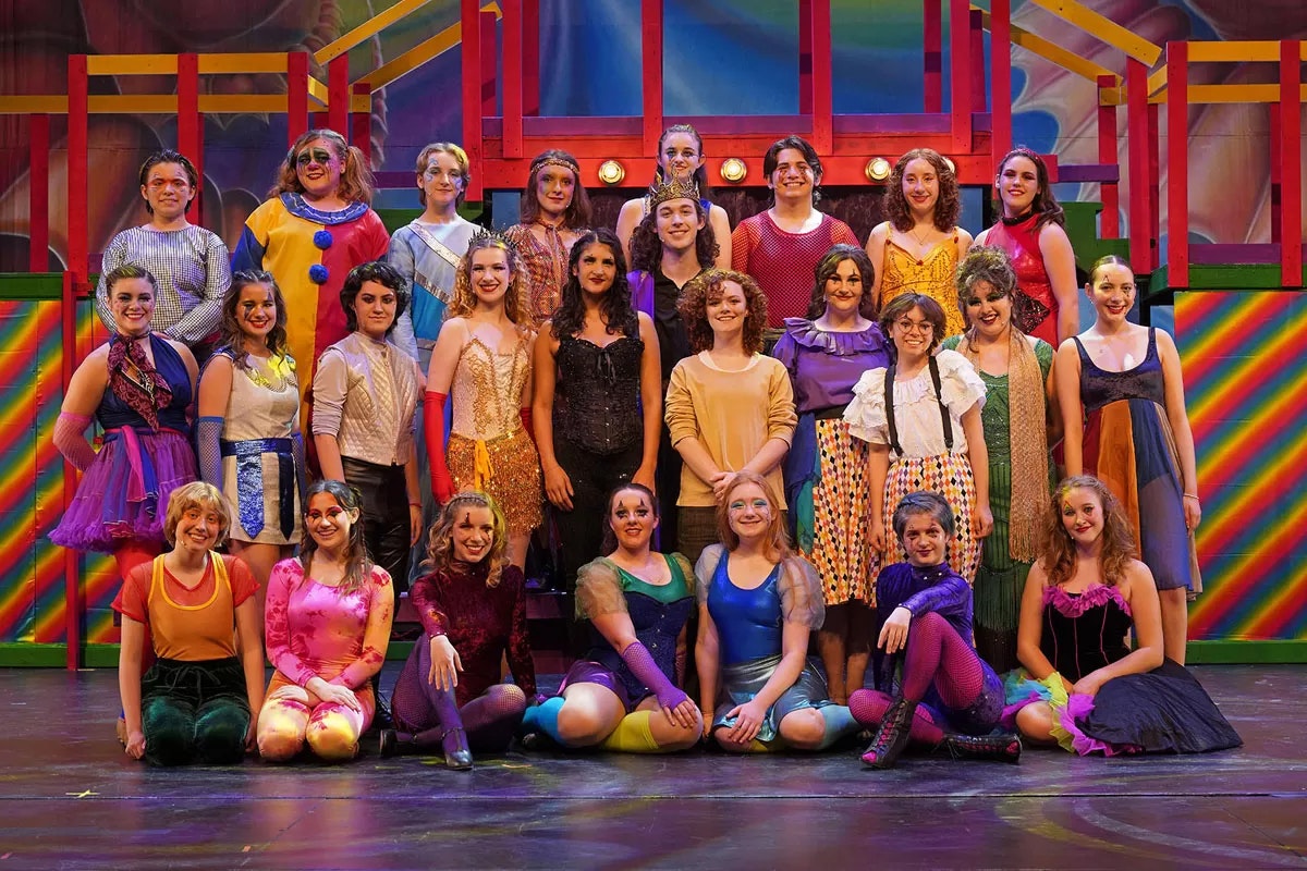 A group of people in colorful costumes posed on stage, in front of a vibrant background.