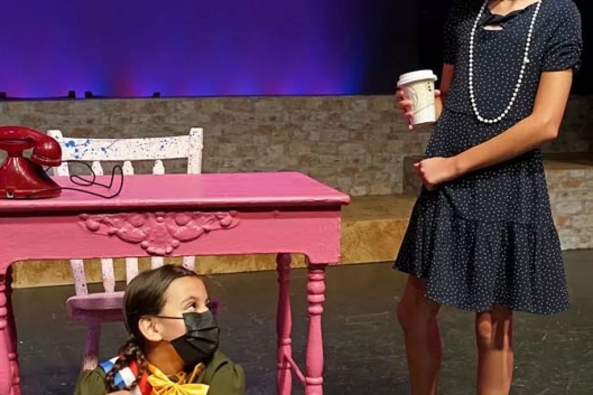 A girl in a polka dot dress holds a drink. Another girl in a mask is under a pink table with a red phone on top.