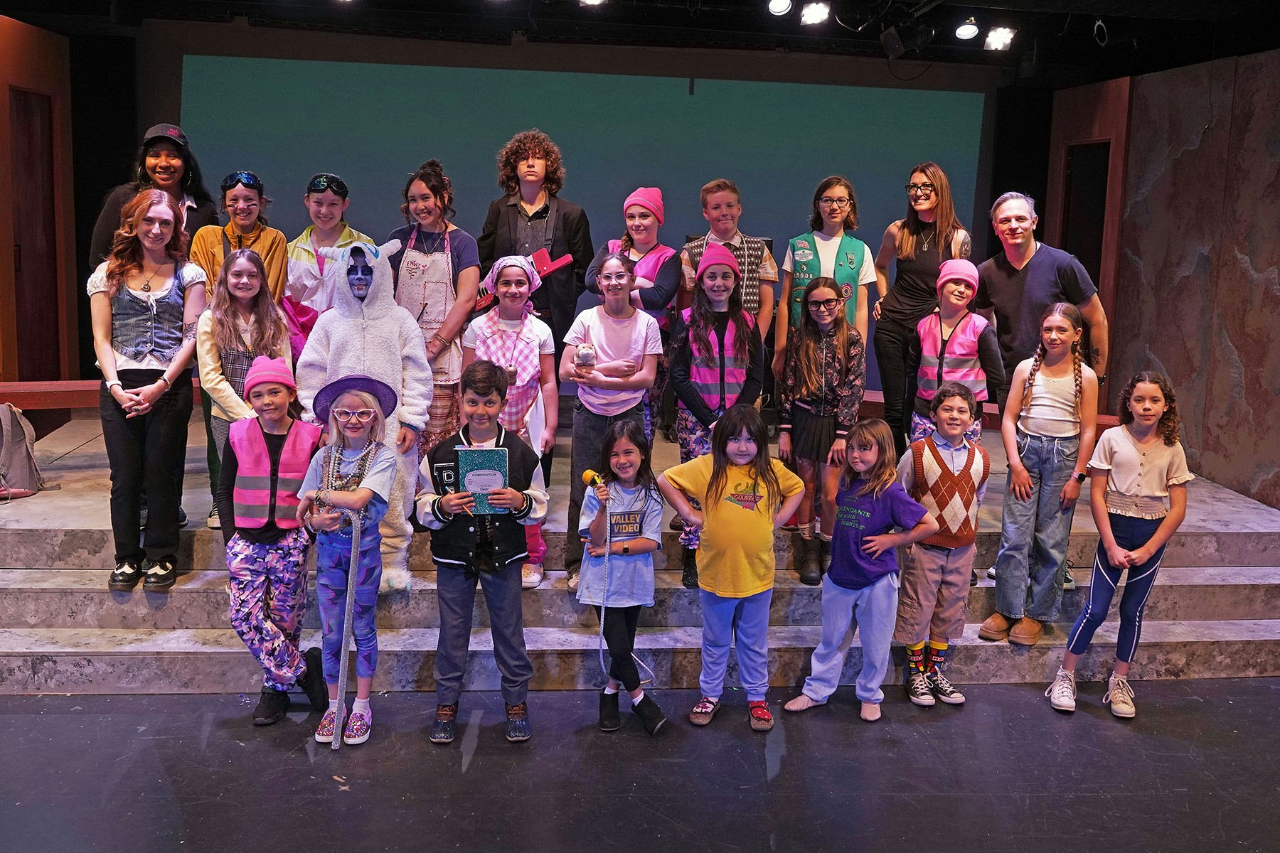 A diverse group of children and adults poses on stage in colorful outfits, likely celebrating a theater performance or event.