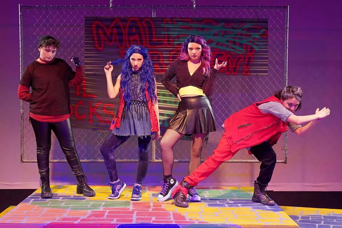 Four people pose on a colorful stage with a chain-link fence backdrop and graffiti.