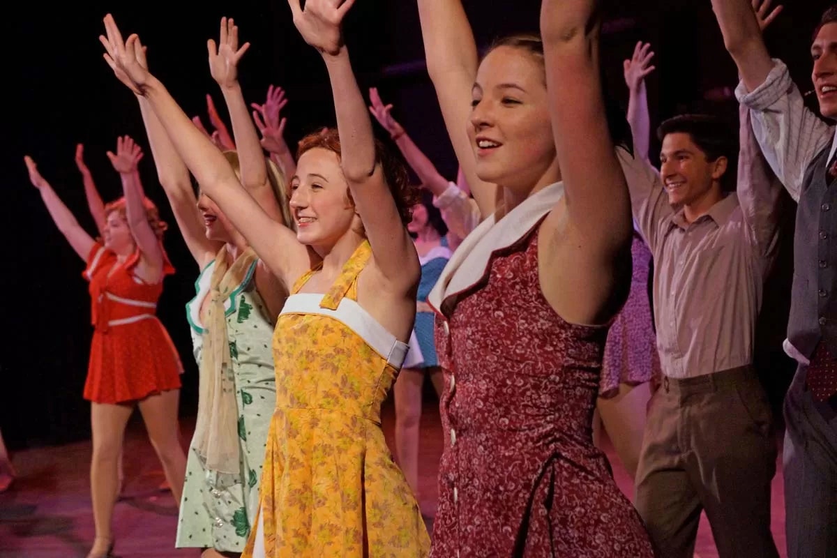 A group of performers on stage in colorful costumes, smiling with arms raised.
