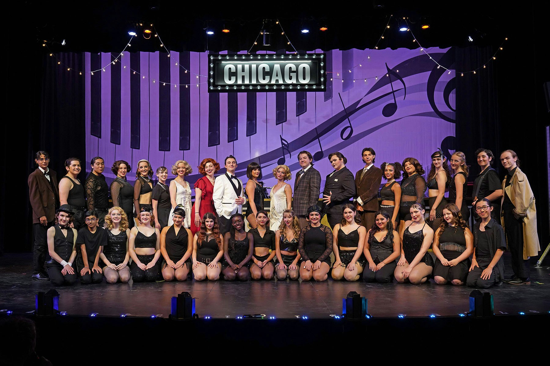 A large group of performers poses on stage for a production of "Chicago," featuring a musical-themed backdrop.