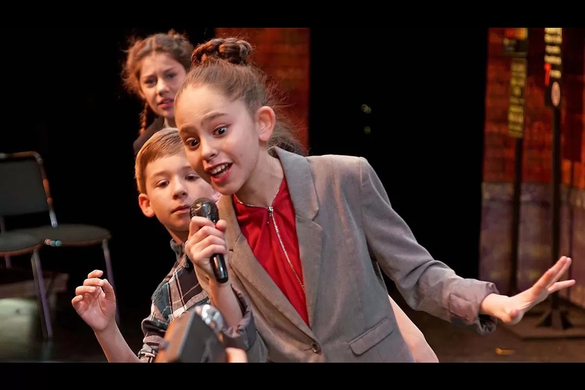 Kids performing on stage; a girl in a suit sings into a microphone, others watch.