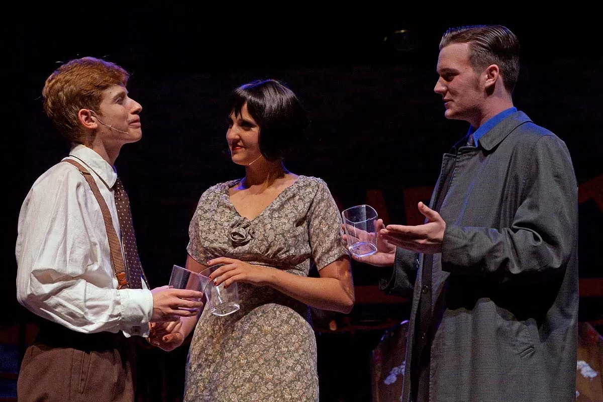 Three actors in costume, holding glasses, engaged in a conversation on stage.
