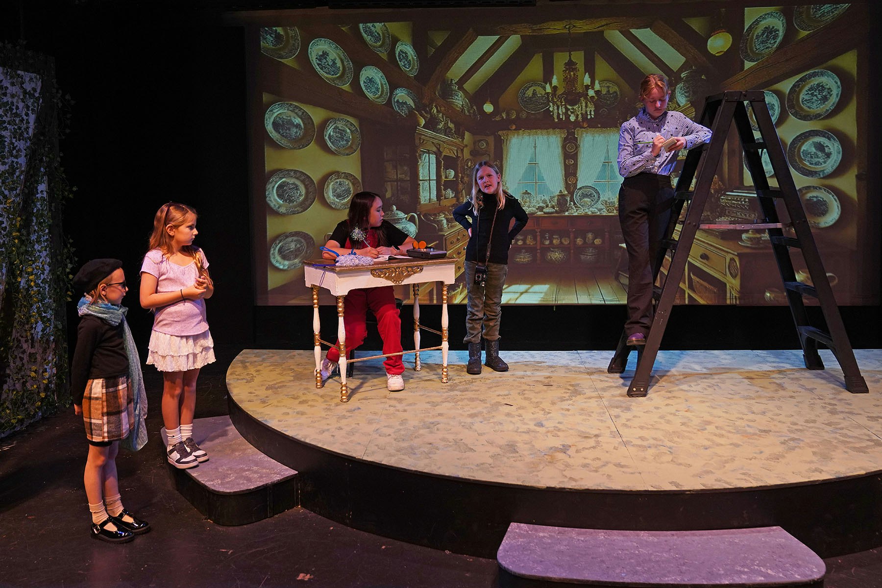 Four people on a stage set resembling a cozy interior, with one person on a ladder and others interacting near a small table.
