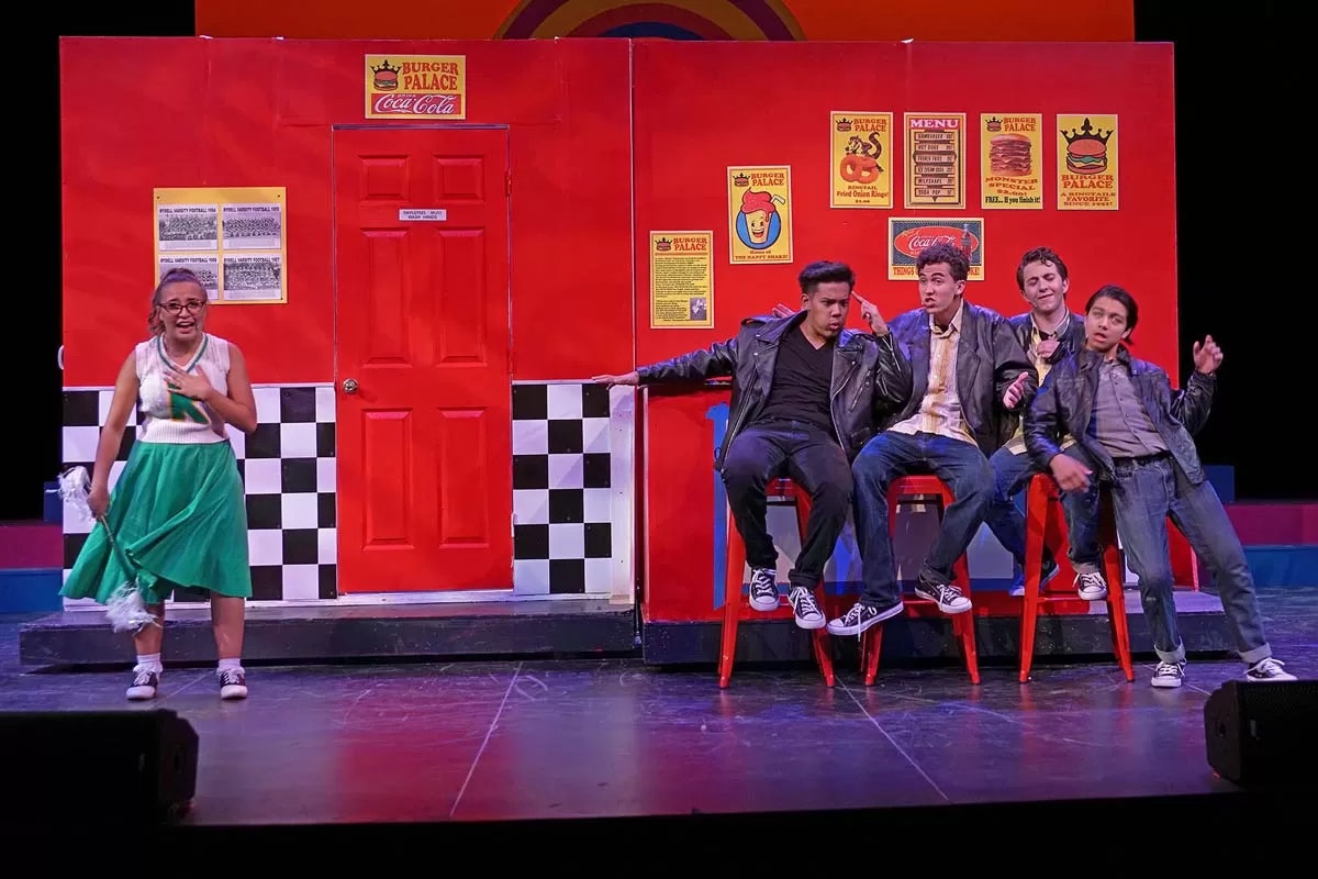 A group of people on stage; one in a dress, others on stools in leather jackets. Bright red setting with vintage posters.