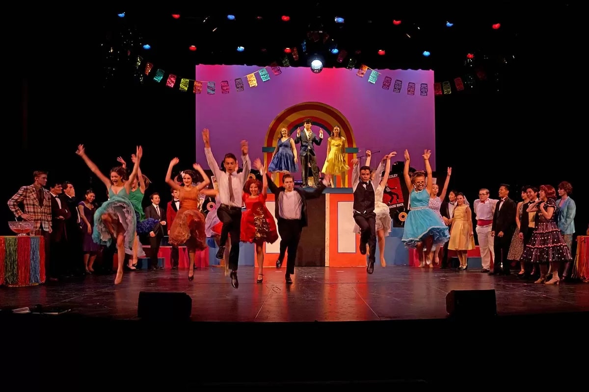A lively stage performance with dancers in colorful costumes mid-jump, surrounded by other performers and vibrant decorations.