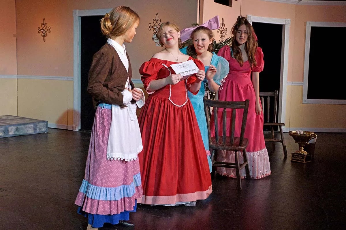 Four women in period costumes interacting on a stage, one holding a letter.