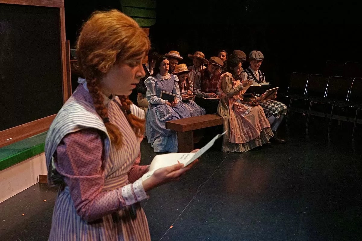 A scene from a play with people dressed in vintage clothing, sitting and holding papers or books.