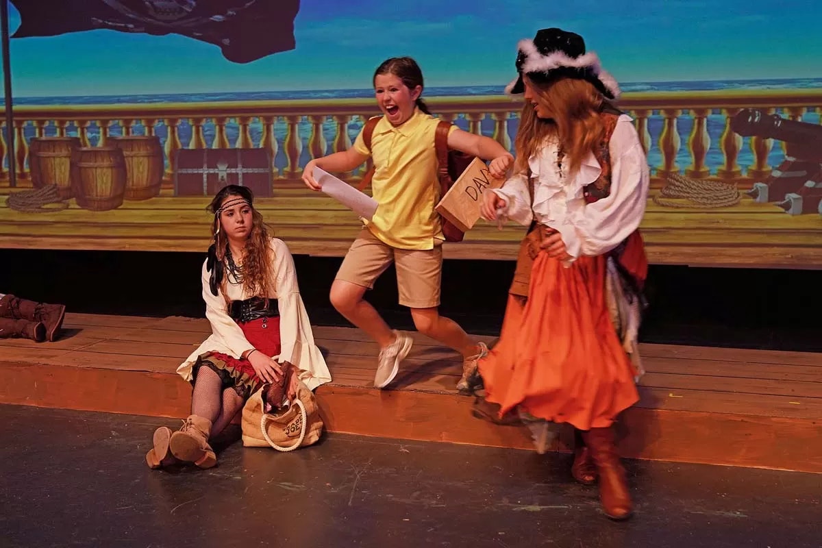 Three people on stage in pirate-themed costumes; one is energetically running, holding paper and a box.