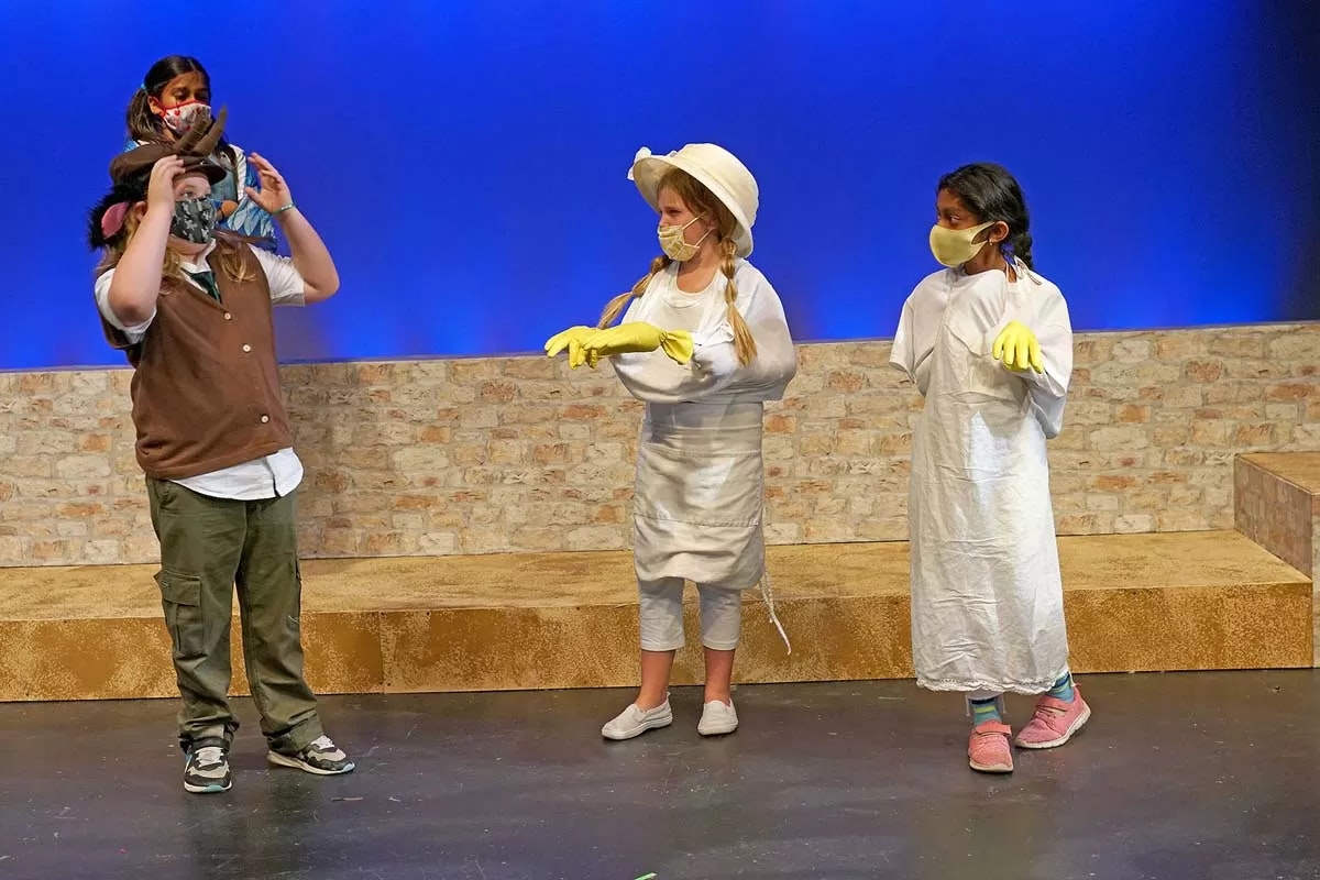 Kids in costume with masks and gloves on a stage set with a blue background and brick wall.