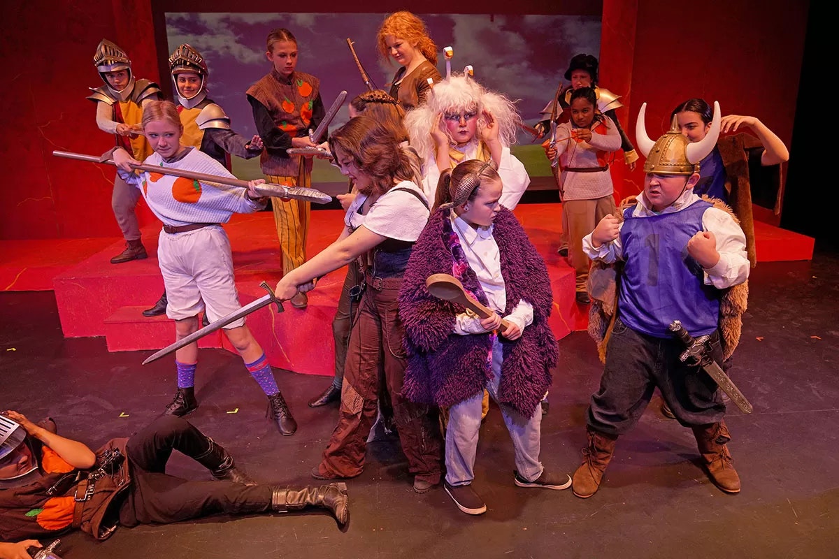 A group of people in colorful costumes on stage with toy swords and props, appearing to perform a theatrical scene.