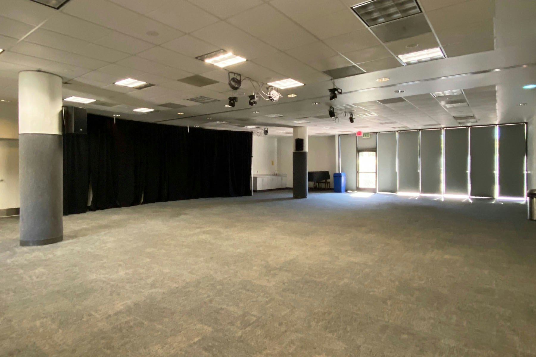 An empty room with carpet, ceiling lights, black curtains, and blinds on windows; features pillars and a blue trash bin.