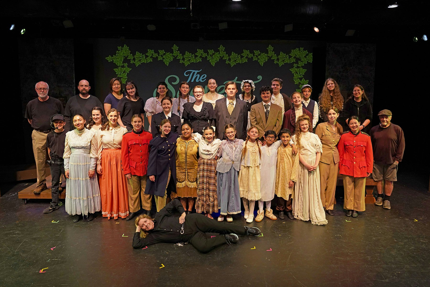 A group of people in period costumes poses on a stage, with one person lying in front. The background is decorated with leaves.