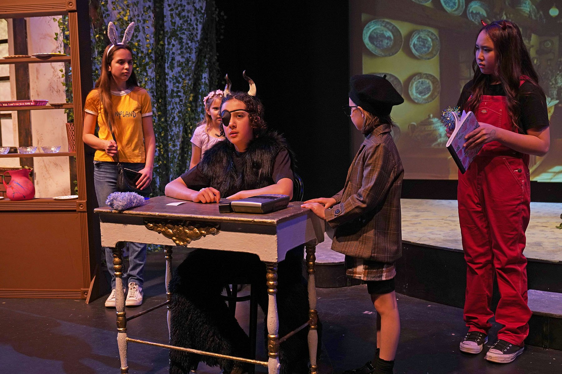 A group of people in costume are performing a play on stage, one sitting at a desk, others standing around.