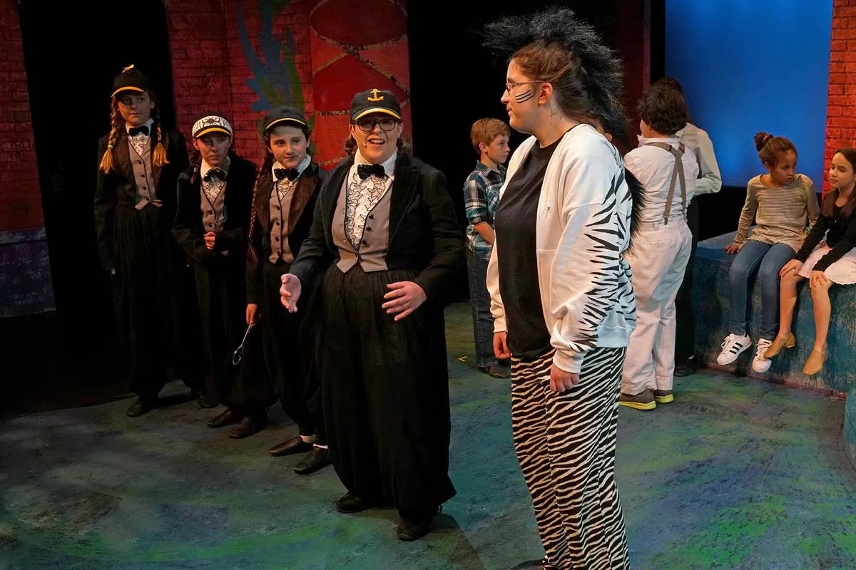 People in costumes on a stage, some in matching suits and hats, another in a zebra-patterned outfit, with others sitting in the background.