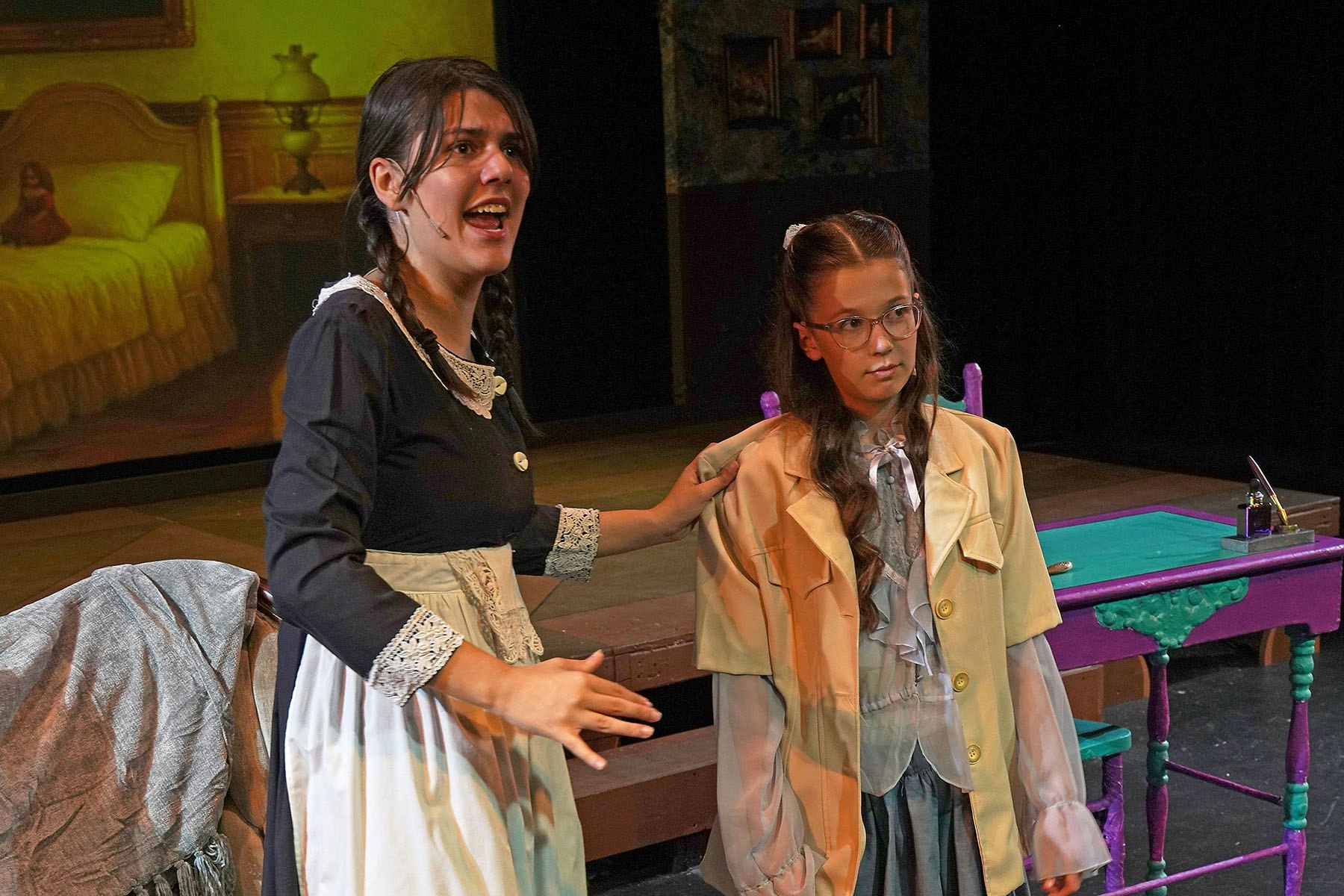 Two actors performing on stage, one gesturing, in a dramatic setting with a background of a bedroom and a desk.